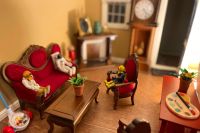 Miniature astronaut and scuba divers sitting on miniature furniture talking with a grandfather clock and fireplace behind them, a paint palette, and toy