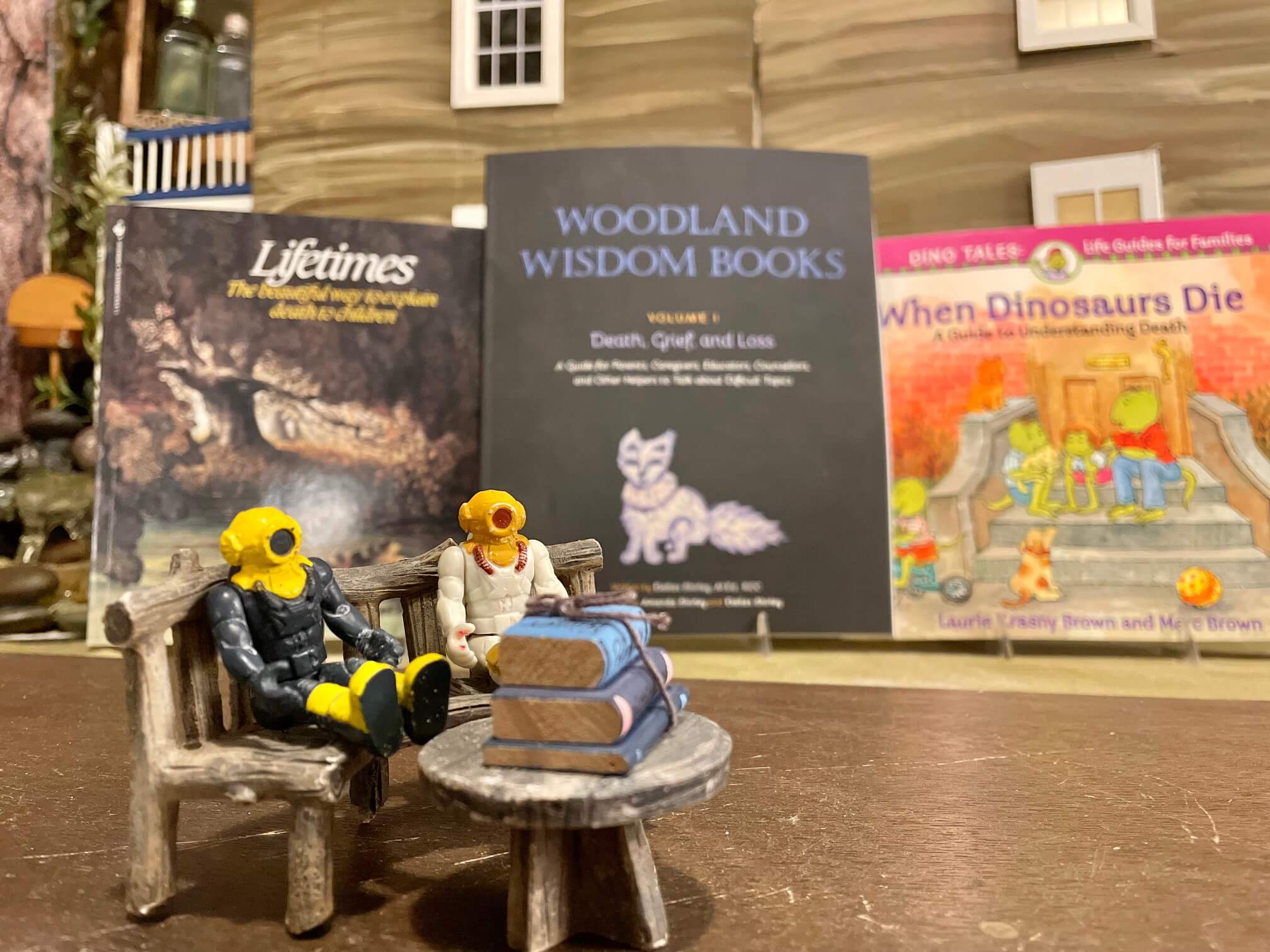 Toys sitting in front of a dollhouse and 3 books focused on death, grief, and loss, and books on the table in front of the toys.