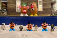 Same toys from last pictures together with 5 Potato Head toys with different facial expressions standing in a sand tray in front of a dollhouse