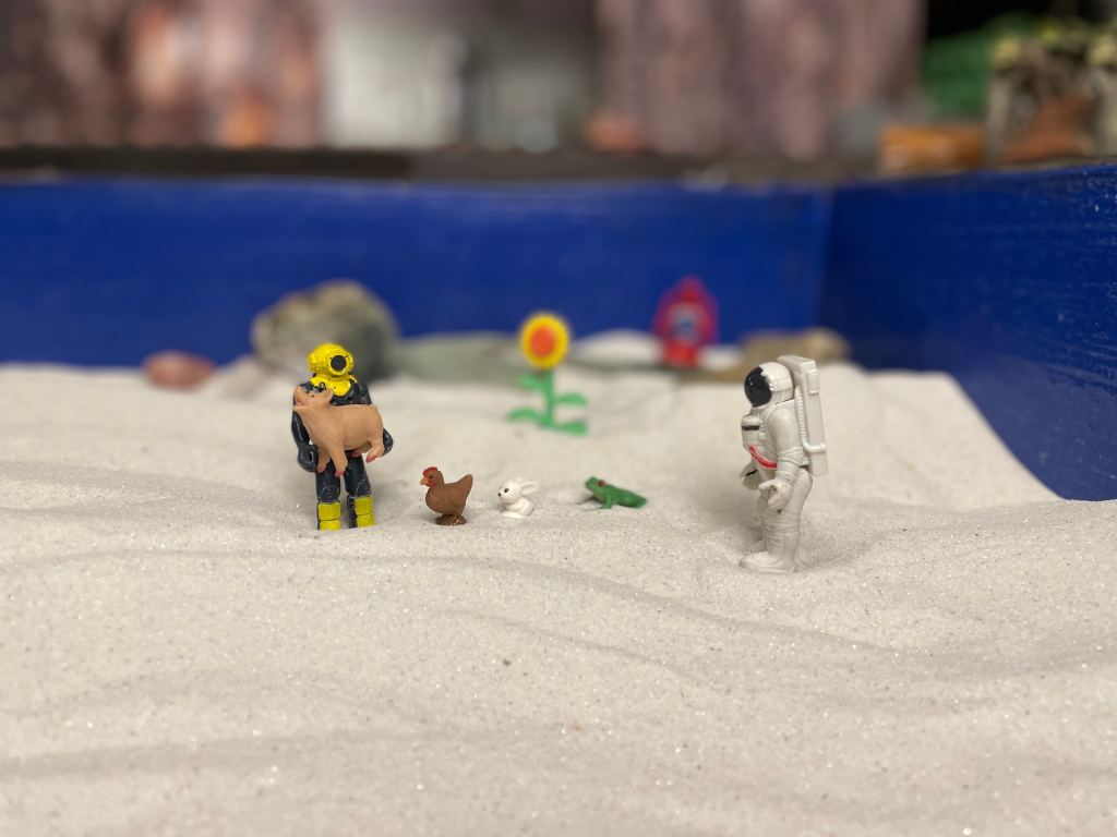 One small toy playing in the sand with small toy animals while another toy is present
