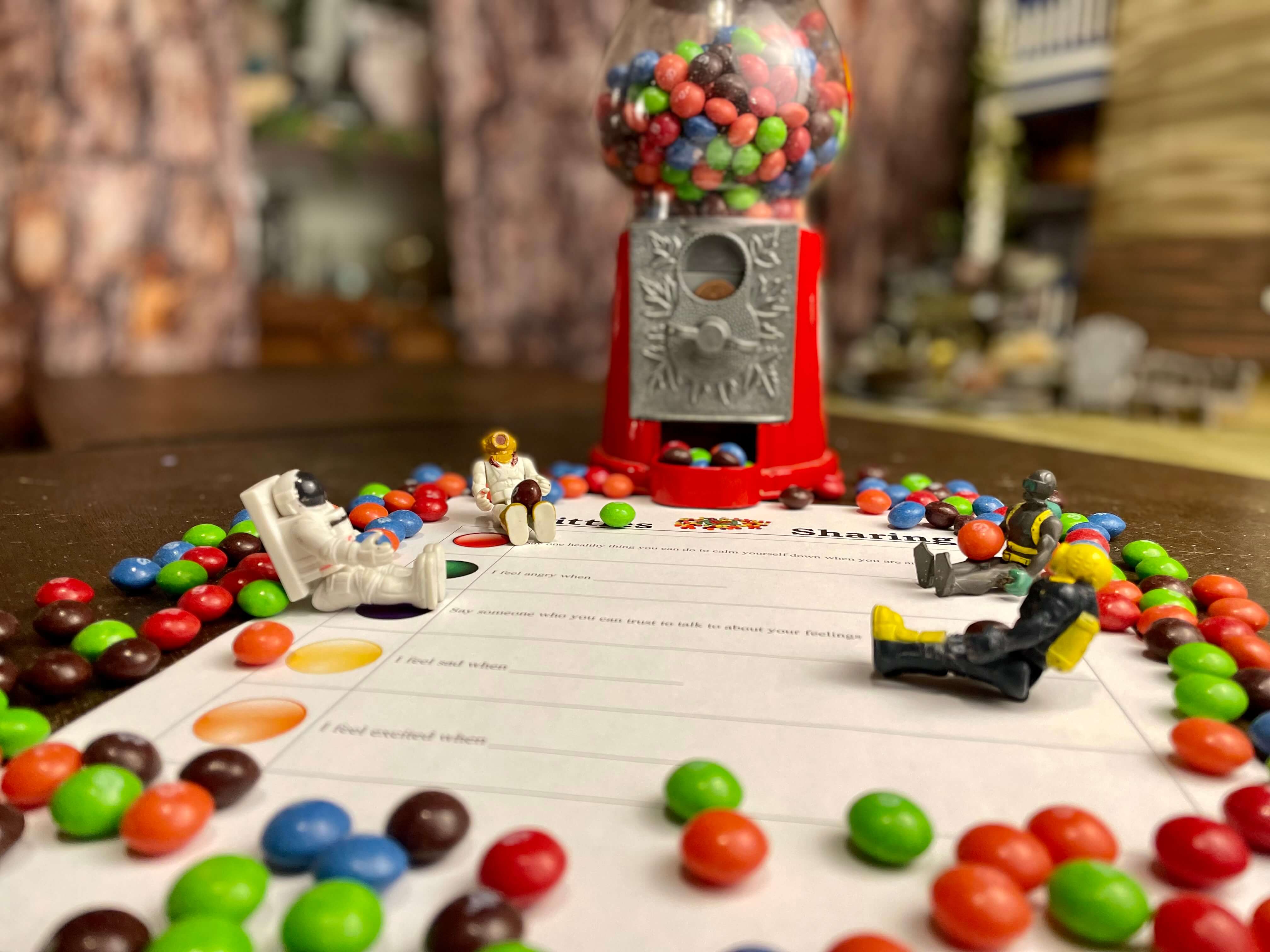 In front of a gumball machine filled with skittles, sits small toys surrounded by skittles on top of a sheet with questions and fill-in-the-blank statements