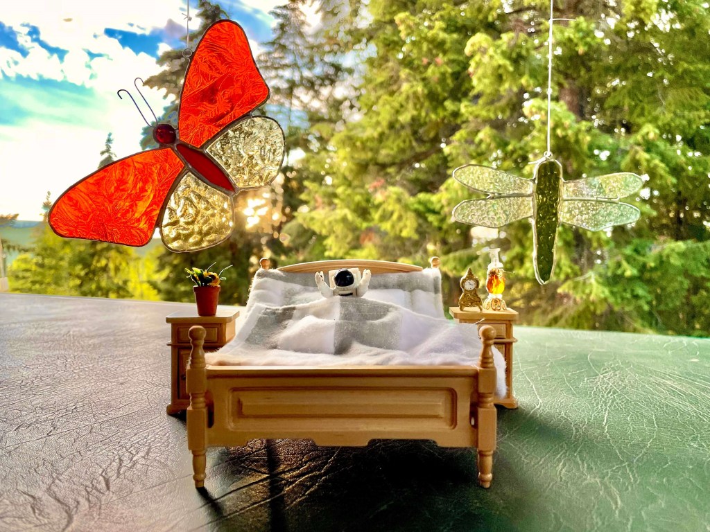 Astronaut toy waking up in bed. Behind them are a dragonfly and butterfly made out of stained glass and a bright sunshine through the trees