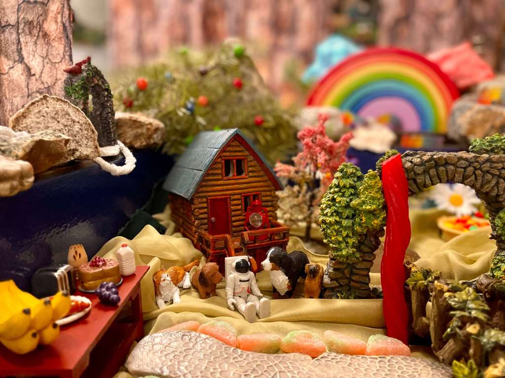 The astronaut miniature in sitting in a forest. They are surrounded by dogs and a cat and sitting in front of a cabin and Wild Berry Skittles trees