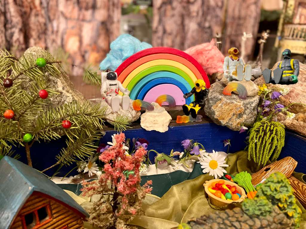 4 miniatures sit on rocks near a rainbow providing shade for cotton candy clouds. There are flowers, fruit, a willow tree, spring tree, and a tree with Skittles