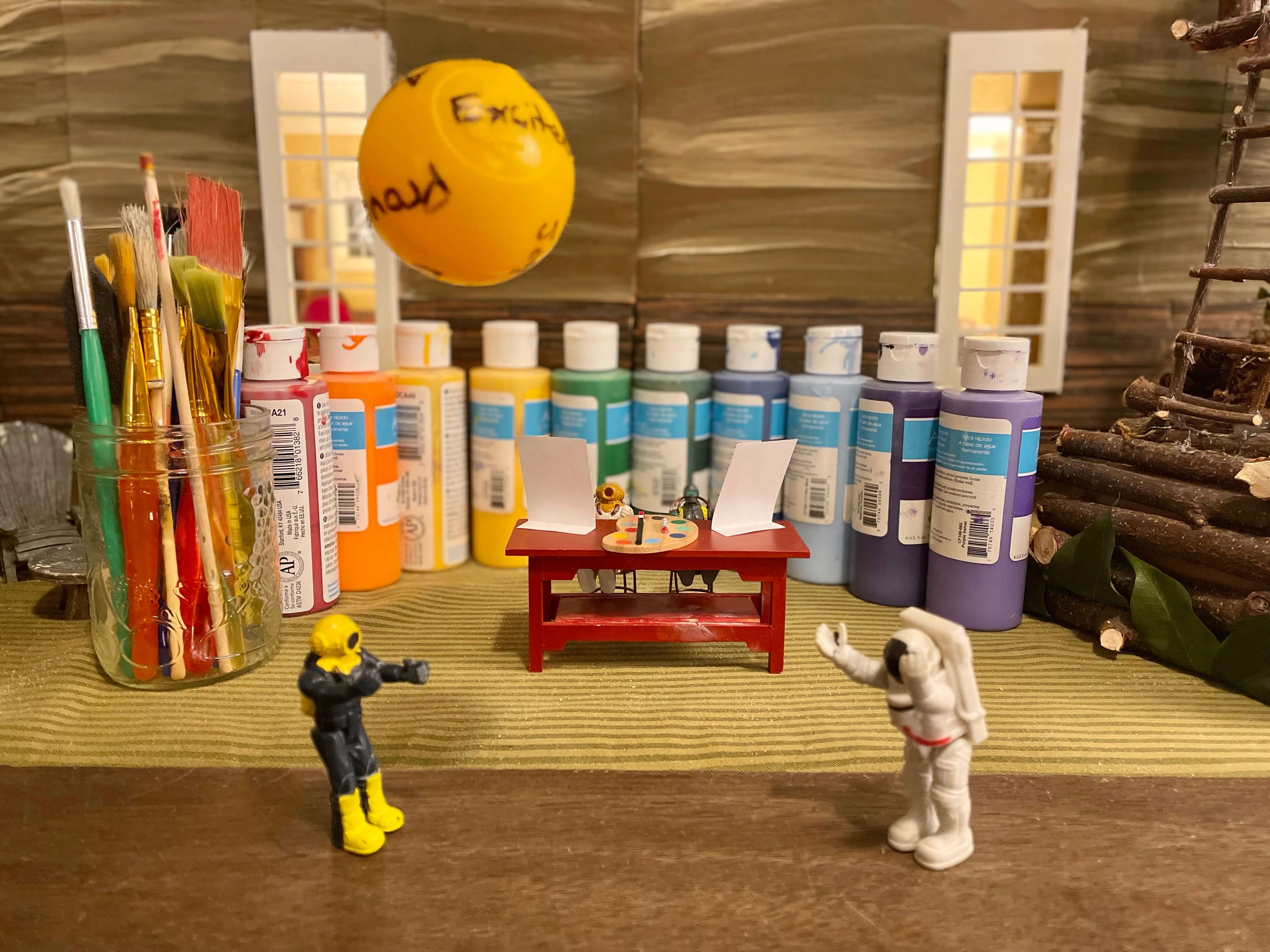 2 miniatures are painting with paint behind them, a table with two easels, a paint brush and palette, while 2 miniatures throw a ball of emotions back and forth