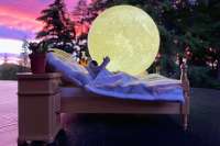 Astronaut toy laying in bed, reaching out, in front of a moon and a pink and purple sunset