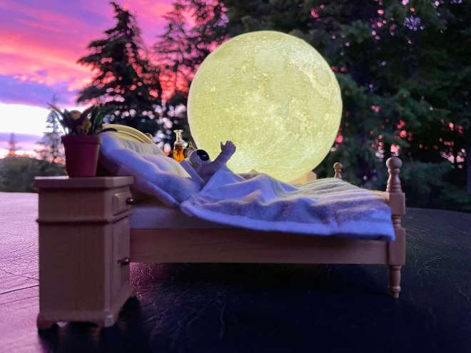 Astronaut toy laying in bed, reaching out, in front of a moon and a pink and purple sunset