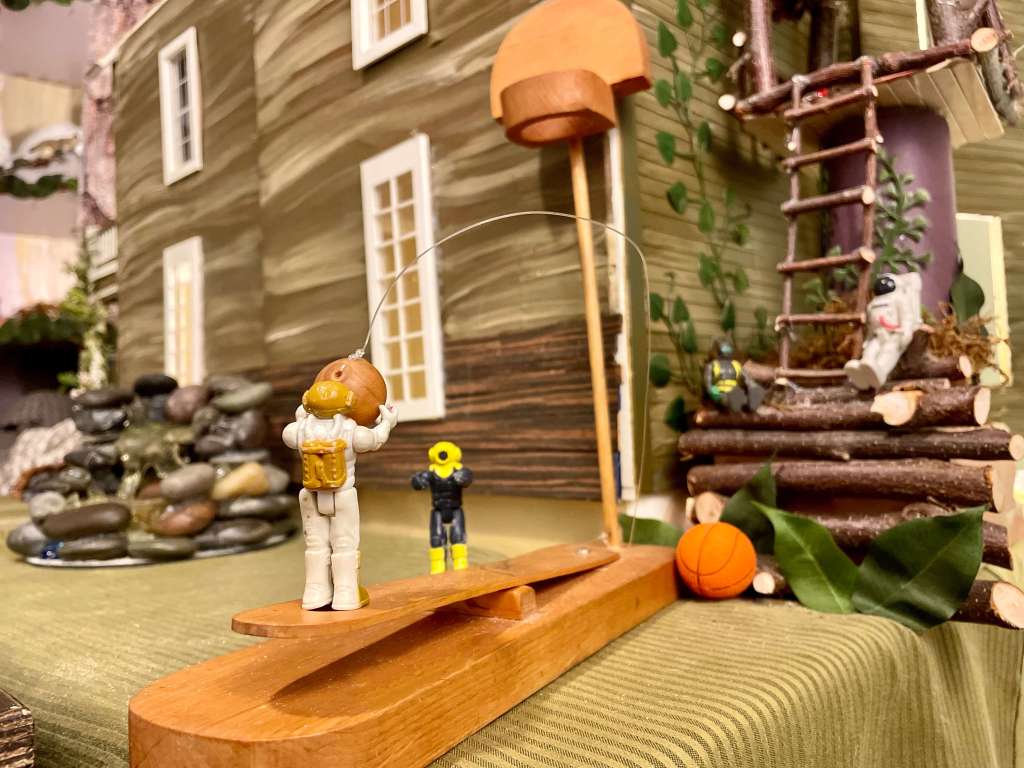 2 toys playing basketball and 2 toys sitting in a treehouse with a dollhouse and fountain in the background