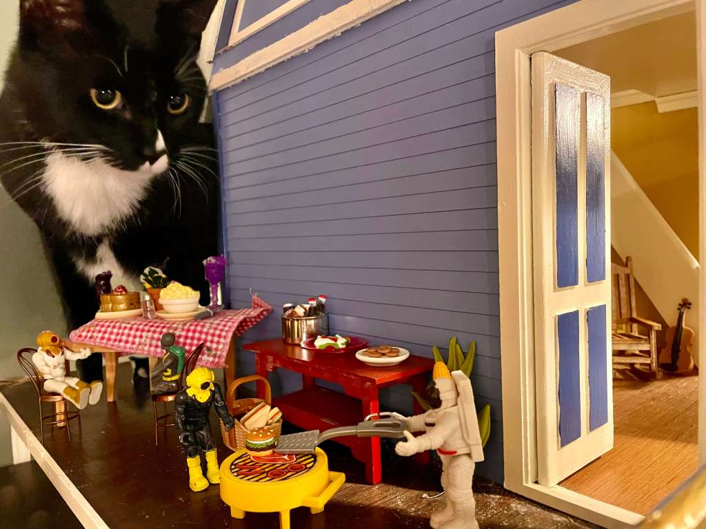 4 toys having a barbeque on a deck, with an empty rocking chair and violin inside the dollhouse and a cat in the back