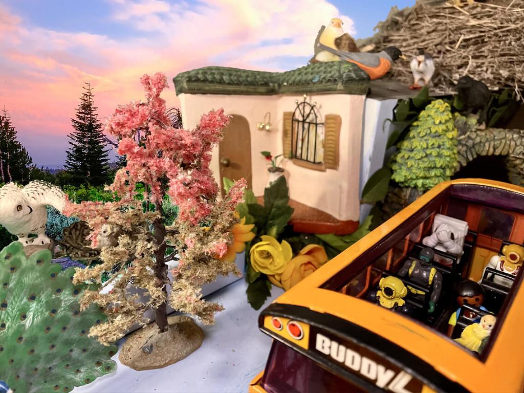 6 toys in a bus with 1 in the back with their hands covering their face driving by birds, a home, trees, and a sunset
