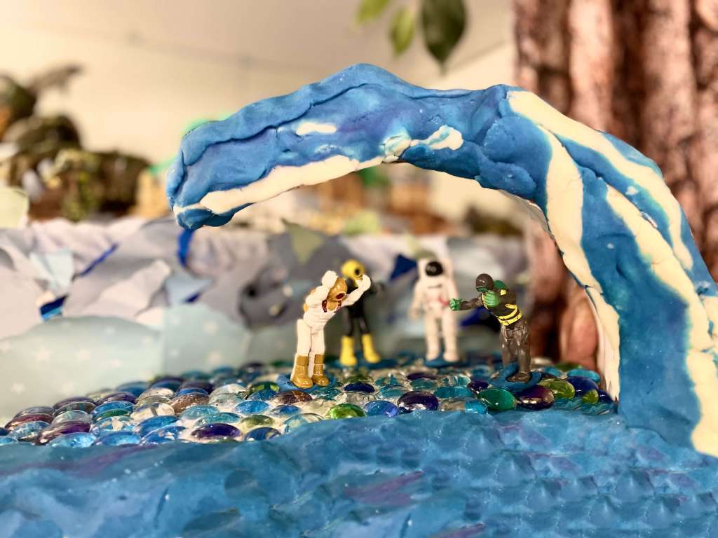 4 toys under a wave of blue and white play dough and standing on water made of blue, green, and clear glass beads