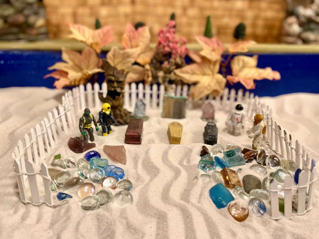 4 toys in a sand tray with 3 coffins and tombstones between them. A fence around them, glass beads, and Fall trees