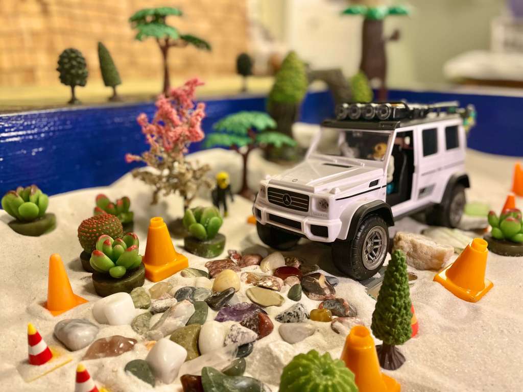 A toy learning how to drive in a sand tray on top of a road made of stones surrounded by orange cones, trees, and bushes
