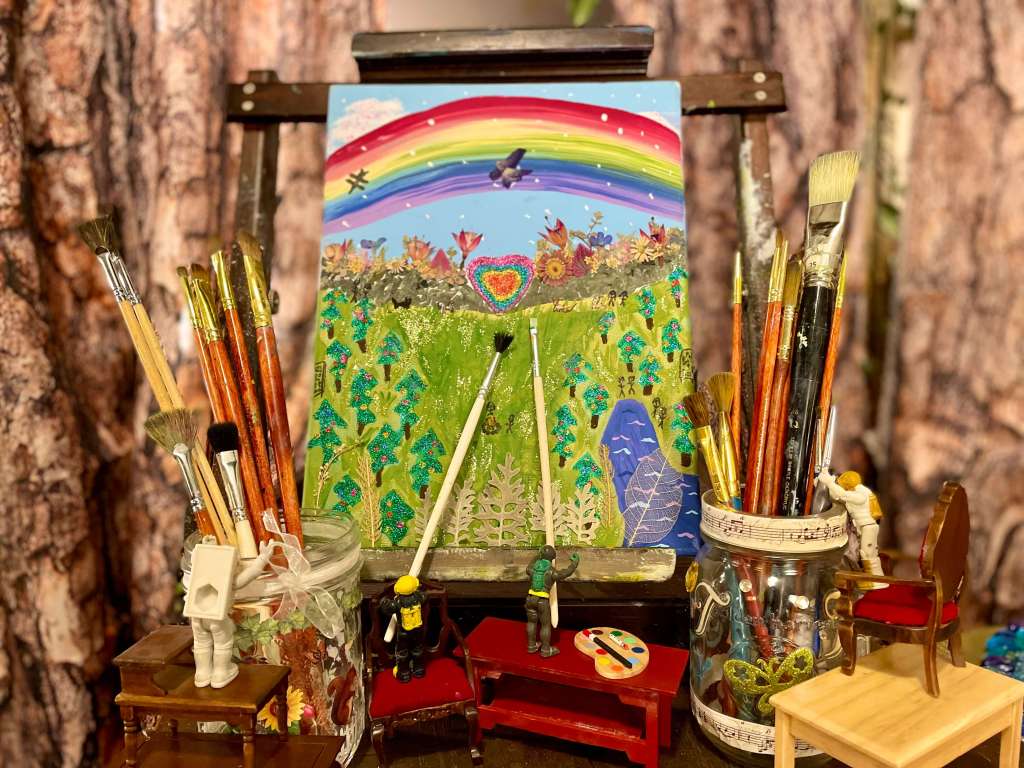 2 toys painting on a big easel with 2 toys standing on miniature furniture trying to reach paint brushes out of mason jars
