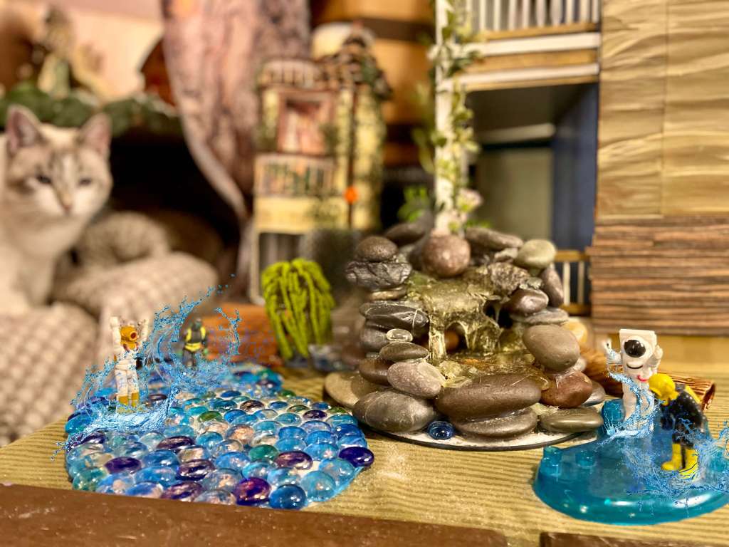 4 toys jumping into puddles with water splashing. A fountain in the middle with a tree, house, castle, and a cat in the back