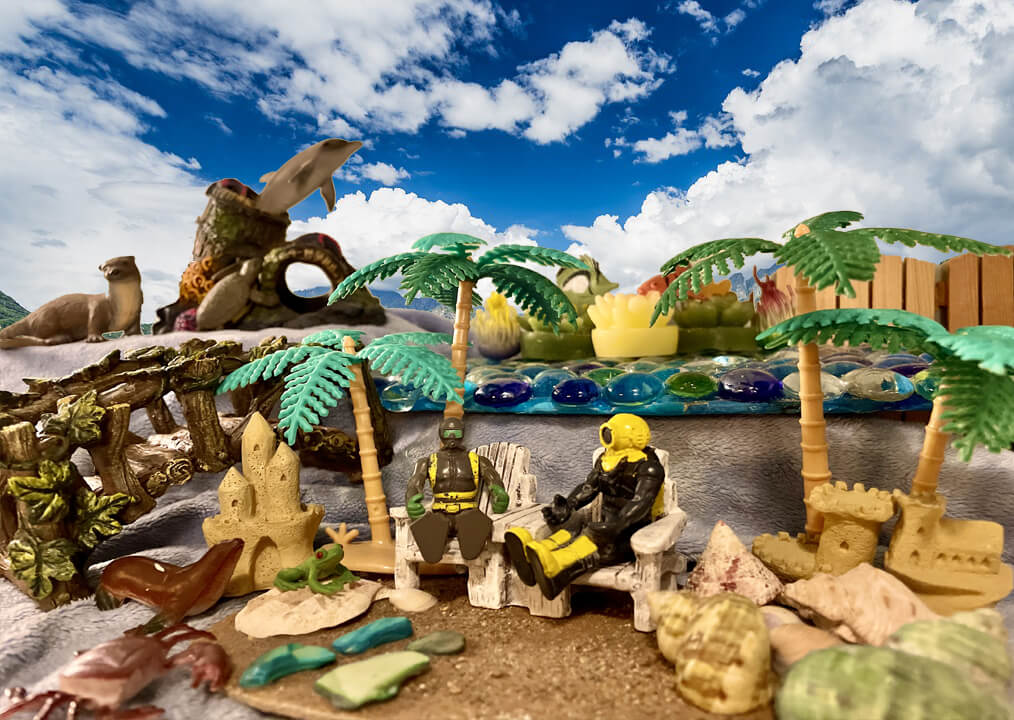 2 toys sitting in lounge chairs in a beach scene with palm trees, a sealion, an otter, a dolphin, sandcastles, and seashells