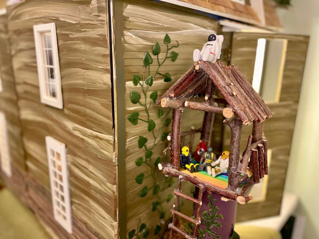 3 toys sitting in a treehouse and 1 toy sitting on the roof of the treehouse all in front of a dollhouse
