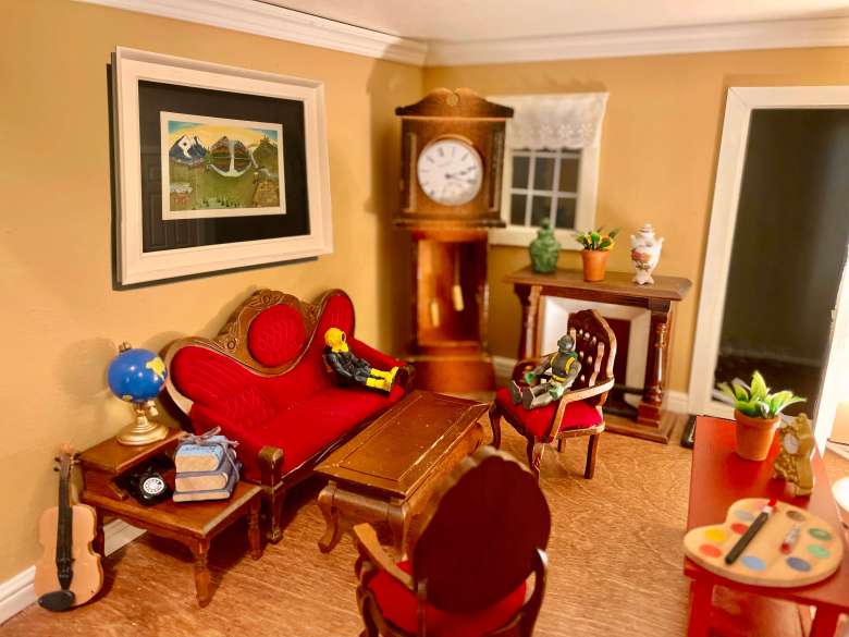 Two toys on couches talking. Behind them is a grandfather clock and a mantle with plants on it. On other tables there are books, a globe, a violin, and a paint palate.